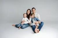 Family of four sitting together in a minimalist studio with light gray background, coordinated neutral outfits and joyful expressions captured by Kaitlyn Dawn Photography, professional family portrait photographer.