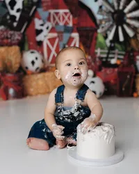 First birthday portrait of a baby boy in denim overalls, sitting in front of a red barn backdrop with cake in hand. Photograph by Yucaipa cake smash photographer Kaitlyn Dawn Photography.