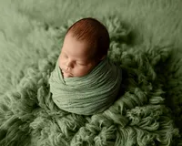 Sleeping newborn baby boy wrapped in a soft green swaddle and posed on a sage green backdrop during a studio newborn photography session by Inland Empire newborn photographer Kaitlyn Dawn Photography.
