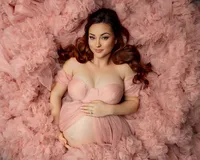 Pregnant woman with red hair lying in layers of a blush pink ruffled maternity gown while gently holding her baby bump during a close-up studio maternity portrait photographed by Inland Empire maternity photographer Kaitlyn Dawn Photography.
