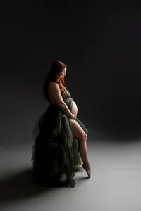 Fine art maternity portrait of a pregnant woman in a dark green gown standing in profile against a dramatic studio background by Kaitlyn Dawn Photography.