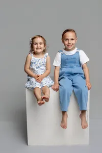 Brother and sister sitting on a white studio cube in a neutral gray studio, coordinated blue outfits and smiling expressions captured by Kaitlyn Dawn Photography, professional children’s and sibling portrait photographer.