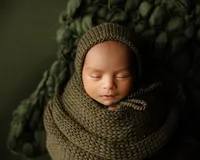 Sleeping newborn baby wrapped in a cozy olive knit swaddle and bonnet on a dark green textured backdrop during a studio newborn photography session by Kaitlyn Dawn Photography.