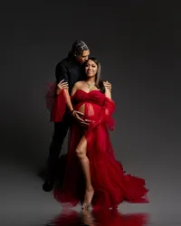 Expecting couple posing together during a dramatic studio maternity photoshoot with mother wearing a flowing red tulle maternity gown while father embraces her and holds her baby bump, photographed by Kaitlyn Dawn Photography.