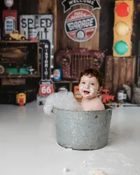 Laughing baby in a bubble bath with frosting on his face, surrounded by retro signs and a rustic truck backdrop. Photograph by Yucaipa cake smash photographer Kaitlyn Dawn Photography.
