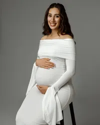Smiling pregnant mom seated on a stool in a white gown, holding her belly during a maternity session by Kaitlyn Dawn Photography in the Inland Empire.