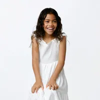 Young girl in a white dress sitting against a white studio backdrop, smiling naturally during a classic child portrait session by Kaitlyn Dawn Photography, professional children’s photographer.