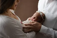 Close-up lifestyle portrait of a newborn baby sleeping in their parents’ arms while wearing a delicate headband during an intimate newborn photography session by Kaitlyn Dawn Photography.