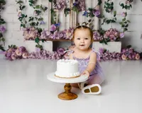 Baby in lavender dress holding a white cake during a cake smash session with purple floral backdrop. Photograph by Yucaipa cake smash photographer Kaitlyn Dawn Photography.