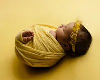 Sleeping newborn baby girl wrapped in a mustard yellow swaddle and wearing a floral headband on a yellow backdrop during a studio newborn photography session.