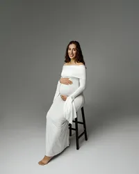 Pregnant woman smiling while sitting on a stool in a long white dress, gently holding her baby bump during an Inland Empire maternity photoshoot with Kaitlyn Dawn Photography.