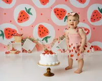 Baby standing beside cake in strawberry dress with watermelon-themed backdrop. Photograph by Yucaipa cake smash photographer Kaitlyn Dawn Photography.