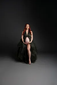 Expecting mother with long red hair wearing an elegant dark green maternity gown while smiling softly and cradling her baby bump during a fine art studio maternity photography session on a dark background by Kaitlyn Dawn Photography.