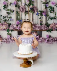 Smiling baby in a lavender “one” dress during a cake smash with purple flowers in the background. Photograph by Yucaipa cake smash photographer Kaitlyn Dawn Photography.