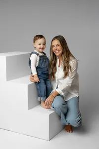 Mother sitting beside her toddler son on modern white studio blocks against a soft gray backdrop, smiling during a timeless studio family portrait session by Kaitlyn Dawn Photography.