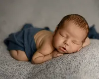 Peacefully sleeping newborn baby boy posed on a textured gray blanket with hands tucked under chin during a professional newborn photography session by Inland Empire newborn photographer Kaitlyn Dawn Photography.