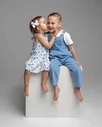 Two siblings sitting on a white studio cube, little girl kissing her brother in coordinated blue outfits, photographed by Kaitlyn Dawn Photography, children’s and sibling portrait photographer.