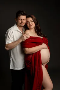 Expecting couple embracing during a studio maternity photoshoot with mother in a red gown photographed by Kaitlyn Dawn Photography.