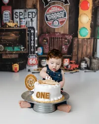 Baby boy in denim overalls digging into a white cake topped with a gold “ONE” and tire-themed decoration, set in a vintage garage backdrop with Route 66 details. Photograph by Yucaipa cake smash photographer Kaitlyn Dawn Photography.
