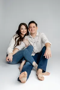 Young couple sitting barefoot on the studio floor against a soft gray backdrop, relaxed and smiling during a natural lifestyle portrait session by Kaitlyn Dawn Photography, professional maternity and family photographer.