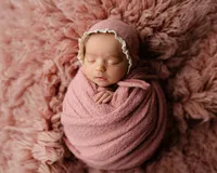 Sleeping newborn baby girl wrapped in a dusty rose swaddle and bonnet on a fluffy pink blanket during a studio newborn photography session by Kaitlyn Dawn Photography in Inland Empire.