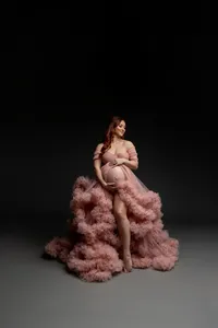 Expecting mother with long red hair wearing an elegant blush pink maternity gown, softly looking down while cradling her baby bump during a fine art studio maternity photoshoot on a dark background by Kaitlyn Dawn Photography.
