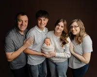 Family portrait with parents and siblings smiling while holding their newborn baby wrapped in a soft neutral blanket during a studio newborn photography session photographed by Kaitlyn Dawn Photography.
