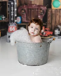 Baby boy with curly hair smiling in a galvanized tub filled with bubbles, foam on his face, set against a vintage garage-themed backdrop with Route 66 signs. Photograph by Yucaipa cake smash photographer Kaitlyn Dawn Photography.
