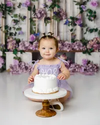 Baby in a lavender “one” dress sitting with a white cake in front of a floral purple backdrop. Photograph by Yucaipa cake smash photographer Kaitlyn Dawn Photography.