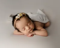 Sleeping newborn baby girl posed with hands under chin and wearing a floral headband on a soft cream backdrop during a studio newborn photography session by Inland Empire newborn photographer Kaitlyn Dawn Photography.