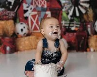 First birthday cake smash featuring a baby boy grinning while covered in cake and frosting, with a barn and hay scene behind him. Photograph by Yucaipa cake smash photographer Kaitlyn Dawn Photography.