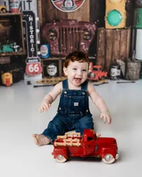 Smiling baby boy in denim overalls playing with a red toy truck in a garage-themed cake smash setup. Photograph by Yucaipa cake smash photographer Kaitlyn Dawn Photography.