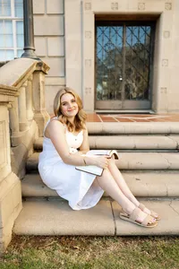 High school senior girl sitting on outdoor stone steps holding a book in a soft natural light setting, senior portrait by Kaitlyn Dawn Photography.