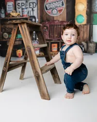 Happy one-year-old boy posing with a small wooden ladder in front of a vintage truck-themed cake smash set. Photograph by Yucaipa cake smash photographer Kaitlyn Dawn Photography.