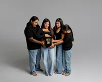 Family gathered around an expecting mom holding a “Native Baby” onesie over her belly during a maternity photoshoot in Yucaipa by Kaitlyn Dawn Photography.