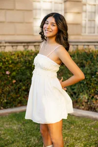 High school senior girl in a white dress standing outdoors near a garden during golden hour, senior portrait by Kaitlyn Dawn Photography.