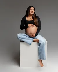 Pregnant mom sitting on a studio block in casual attire, softly holding her belly and looking off-camera during a maternity session in the Inland Empire by Kaitlyn Dawn Photography.