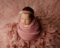 Sleeping newborn baby girl wrapped in a dusty rose swaddle and wearing a delicate headband while posed on a soft pink fur blanket during a studio newborn photography session by Kaitlyn Dawn Photography.