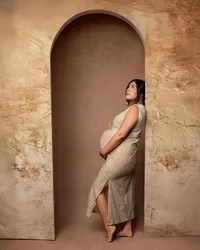 Expecting mom standing in an arched backdrop wearing a beige dress, highlighting her pregnancy during a maternity session by Kaitlyn Dawn Photography in Yucaipa.