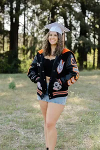 High school graduate wearing a cap and letterman jacket standing in a grassy field, graduation portrait captured by Kaitlyn Dawn Photography.