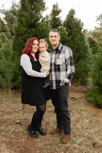 Parents standing together with their baby in a wooded outdoor setting, coordinated neutral outfits captured by Kaitlyn Dawn Photography, lifestyle family portrait photographer.