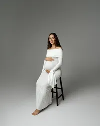 Pregnant woman sitting on a stool in a long white dress, gently holding her baby bump during an Inland Empire maternity photoshoot with Kaitlyn Dawn Photography.