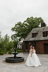 Two brides in white wedding gowns standing near a stone fountain in front of the historic Tudor House at Portage Lakes in Akron, Ohio.