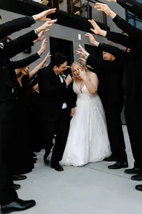 Bride and groom laugh and celebrate walking through cheering wedding party in black formal attire.