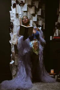 A woman in a voluminous lavender gown reads a black book beside a wall covered in pages and flowers, creating a dramatic, dreamlike mood.