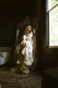 A woman in a flowing white dress and fur wrap poses in an ornate mirror, lit by a window; the dark room feels elegant and mysterious.