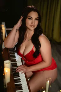 Confident woman in red lingerie sits at a piano, touching her hair and keys, surrounded by glowing candles in a warm, intimate setting.