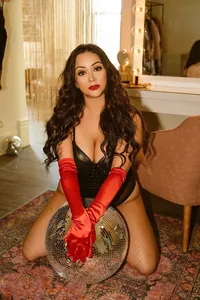 A glamorous woman with long dark curls kneels on a patterned rug in a black outfit and red satin gloves, holding a mirrored disco ball.