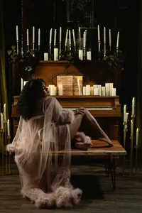 A woman in a sheer, feather-trimmed robe reclines at a candlelit piano, surrounded by candelabras, flowers, and warm, moody shadows.