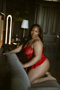 A confident Black woman in red lace lingerie kneels on a velvet chaise, smiling at the camera in a warmly lit, elegant room with a lamp.
